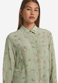 Lys grøn bluse med et blomstermønster med pink og hvide blomster. Klassisk krave, knaplukning foran og lange ærmer. Glat tekstur.