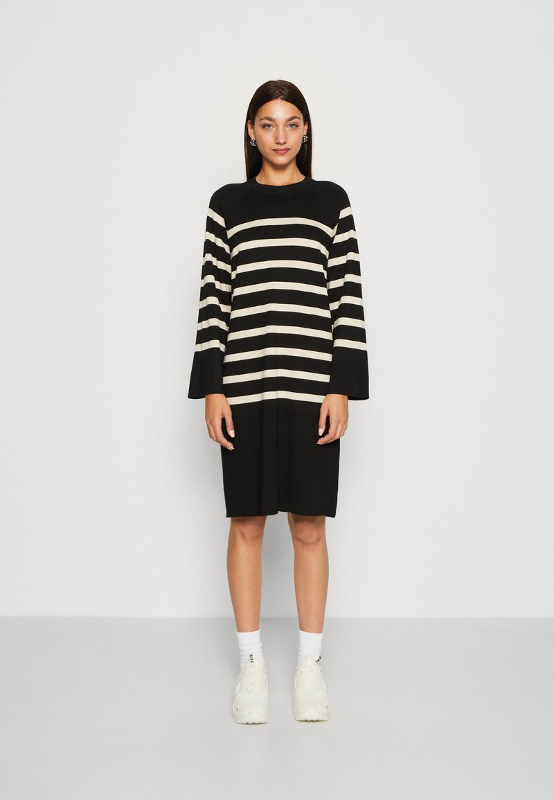 Robe rayée noire et crème avec un col ras du cou et des manches en cloche, réalisée en tissu tricoté, longueur genou, portée avec des baskets blanches et des chaussettes.