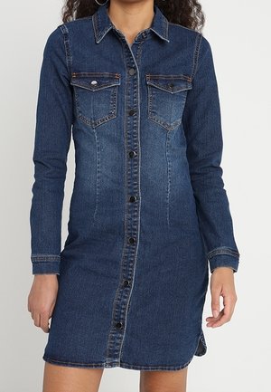 Woman wearing a fitted, long-sleeve, button-up dark blue denim dress with chest pockets and collar.