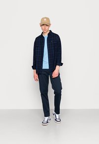 Marineblau karierter Flanellhemd über einem hellblauen T-Shirt, kombiniert mit marineblauen Hosen und weißen Sneakern mit marineblauen Akzenten; beiger Hut.