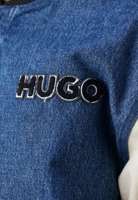 Kurtka dżinsowa z niebieskiego materiału, z czarno-białym logo "HUGO" wyszytym na piersi. Gładka tekstura, klasyczny design.