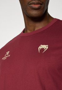 Μπορντό κοντομάνικο μπλουζάκι από μαλακό ύφασμα, με χρυσό λογότυπο και διακριτικό σχέδιο. Στρογγυλή λαιμόκοψη με απαλή υφή.