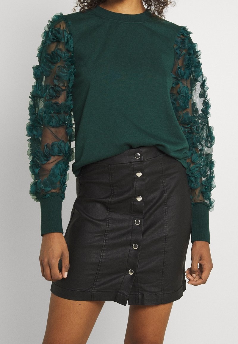 Femme portant un pull vert foncé avec des manches transparentes et volantées, associé à une jupe en cuir noire à boutons.