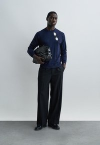 Homme portant un pull marine avec des lunettes de soleil accrochées, un pantalon large noir et des chaussures noires, tenant un sac de sport en cuir noir.