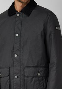 Schwarze Jacke mit einem Steppkragen, Druckknöpfen, zwei Vordertaschen und einem Logopatch am Ärmel. Glatte Textur, hüftlanger Schnitt.