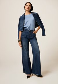 Chaqueta de mezclilla con hombros adornados, combinada con una blusa azul clara y jeans de pierna ancha de cintura alta. Tacones negros de punta.