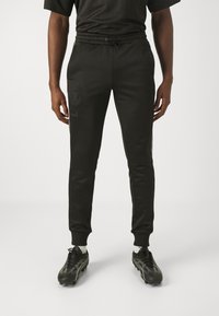 Black jogger pants with elastic waistband, featuring a textured logo on the thigh and tapered ankles. Worn with black cleats and white socks.