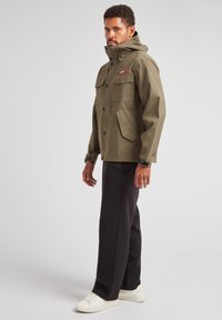 Giacca impermeabile verde oliva con colletto alto, quattro tasche frontali e bottoni a pressione. Abbinata a pantaloni neri e sneakers bianche.