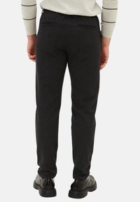 Černé, zúžené kalhoty s hladkou texturou, elastickým pasem a dvěma zadními kapsami. Nošeny s černými botami, pohled zezadu.