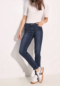 Vaqueros slim-fit azul oscuro con una cintura de altura media, costuras decorativas y dos bolsillos frontales. Combinados con zapatillas blancas y una blusa clara.