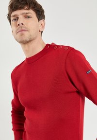 Roter gestrickter Pullover mit rundem Halsausschnitt, geripptem Muster und vier Knopfdetails an der Schulter. Lange Ärmel mit enganliegenden Bündchen.