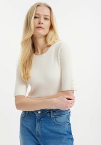 Žebrovaný svetr krémové barvy s krátkými rukávy a knoflíkovými detaily na manžetách, nošený s modrými džíny s vysokým pasem.