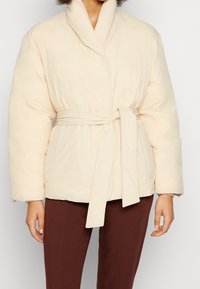 Beige vadderad jacka med skärp, med sjalkrage, långa ärmar och två sidofickor, i mjuk, quiltad textur.