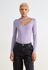 Lilla langermet bodysuit med utskåret utringning og Puma-logo. Brukt med høyt midje sorte bukser, som viser en avslappet passform og gullakenter.