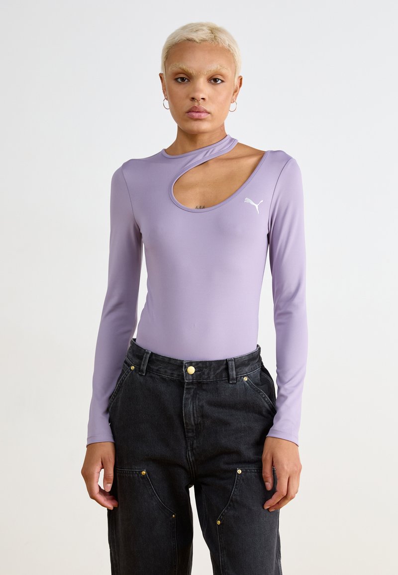 Lilla langermet bodysuit med utskåret utringning og Puma-logo. Brukt med høyt midje sorte bukser, som viser en avslappet passform og gullakenter.