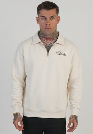 Mand med tatoveringer på nakke og hånd iført en cremefarvet sweatshirt med kvart lynlås og "SikSilk"-logo på brystet samt sorte bukser.