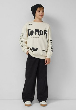 Kremowa bluza z czarnym napisem, luźny krój. Luźne czarne spodnie. Białe trampki z drewnianą podeszwą, czarna czapka typu beanie i swobodna postura.
