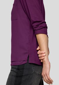 Langes lila Langarmshirt mit rundem Halsausschnitt und Seitenschlitzen, aus weichem Stoff. Verfügt über eine einzelne Tasche auf der linken Seite.