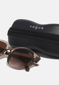 Ochelari de soare translucizi maro cu lentile întunecate și o carcasă neagră cu fermoar, având un logo alb "vogue". Forma lentilelor este supradimensionată și rotundă.