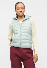 Νεαρή γυναίκα με πλεγμένα μαλλιά που φοράει ανοιχτό μπλε γιλέκο με καπιτονέ σχέδιο πάνω από κρεμ πουλόβερ και βουργουνδί παντελόνι, ποζάροντας με το ένα χέρι στην τσέπη.