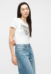 Νέα γυναίκα με μακριά ίσια μαλλιά, φορώντας λευκό μπλουζάκι Guess με στάμπα και ανοιχτό γαλάζιο τζιν ψηλόμεσο, στέκεται μπροστά σε λευκό φόντο.