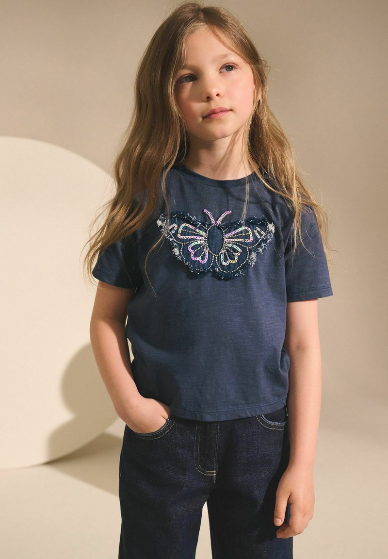 Marinblå t-shirt med broderad fjärilsdesign som har färgglada pärlor och fransade detaljer, ihop med mörka denimjeans.