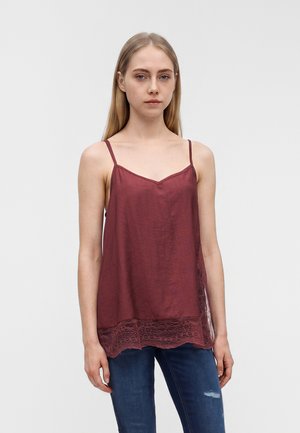 Frau mit glattem blondem Haar, die ein ärmelloses bordeauxfarbenes Spitzenoberteil und dunkelblaue Jeans trägt, vor einem schlichten Hintergrund stehend.