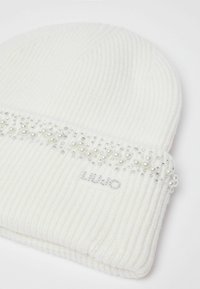 Bonnet en tricot blanc avec texture côtelée, orné de pierres claires et d'accents semblables à des perles. Présente un logo cousu en bas.