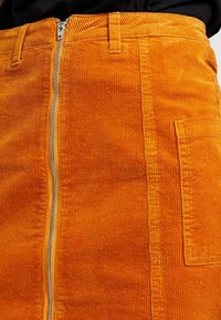 Orange kordkjol med dragkedja framtill, med vertikalt ribbat mönster och en sidoficka. Midjan har bälteshällor.