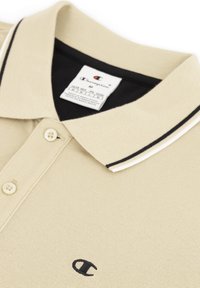 Beige poloshirt van een structuurstof, met een zwart-wit gestreepte kraag en een klein zwart logo op de borst. Beschikt over twee knopen.