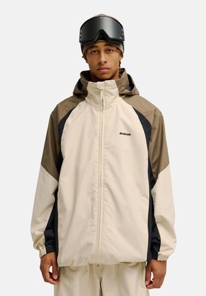 Junger Mann trägt eine beige-braune Skijacke mit schwarzen Akzenten und schwarze Skibrille auf grauem Hintergrund.