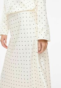 Jupe à pois blancs avec des points noirs, en tissu doux. Elle présente une taille haute et une silhouette fluide, avec des manches longues visibles.