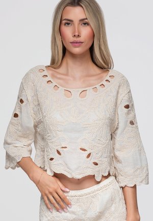 Femme blonde à l'expression neutre portant un haut en dentelle crochet beige avec des détails découpés et un bas taille haute assorti.