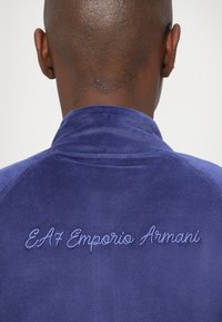 Plava velur košulja s visokim ovratnikom, s izvezenim tekstom "EA7 Emporio Armani" na leđima. Glatka tekstura i prilagođeni dizajn.