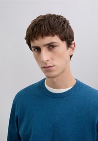 Niebieski sweter wykonany z miękkiego dzianinowego materiału, z okrągłym dekoltem i subtelnym detalem w postaci logo. Tekstura jest gładka i jednolita na całej powierzchni.