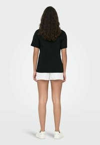 Black cotton t-shirt with short sleeves, shown from the back. Paired with white distressed shorts and white shoes. Smooth texture.