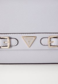 Jasnoszara teksturowana torebka z złotym trójkątnym logo, ozdobiona dwoma złotymi klamrami oraz kontrastującym ciemnym obszyciem na krawędziach.