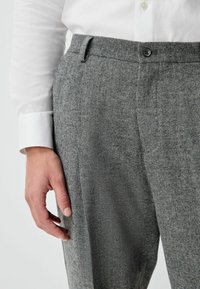 Gray textured trousers with a fine herringbone pattern, featuring a button closure and side pockets, paired with a white shirt sleeve.