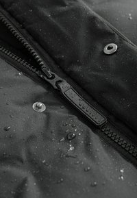 Gros plan d'une fermeture éclair noire mouillée avec des gouttes d'eau sur un tissu résistant à l'eau et deux boutons-pression métalliques.