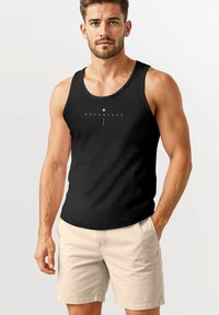 Schwarzes Tanktop mit Rundhalsausschnitt, das weißen Text "NEVERLESS" und ein grafisches Design auf der Vorderseite zeigt. Trägt dazu eine hellbeige Shorts.