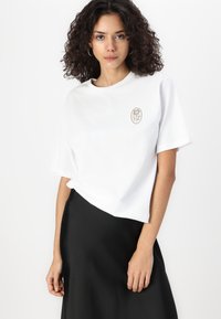 T-shirt blanc en coton à manches courtes, arborant un logo floral beige sur la poitrine. Associé à une jupe en satin noire.