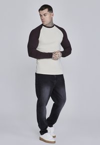 Langærmet, crème skjorte med mørk burgunder raglanærmer, båret med sorte jeans og hvide sneakers. Har et logo på brystet.