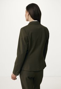 Blazer aderente verde oliva, tessuto liscio, spalle strutturate, polsini a un bottone, pince posteriori per la vestibilità, design minimalista.