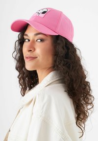 Rosa Baseballmütze mit gesticktem Charakter-Patch, gebogenem Schirm und Baumwollstoff. Leicht schräg getragen mit einer Jacke in neutralen Tönen.