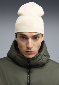 Cappello lavorato a maglia color crema con una vestibilità morbida, caratterizzato da un dettaglio del logo. Abbinato a una giacca a vento verde scuro, con zip frontale e colletto alto.