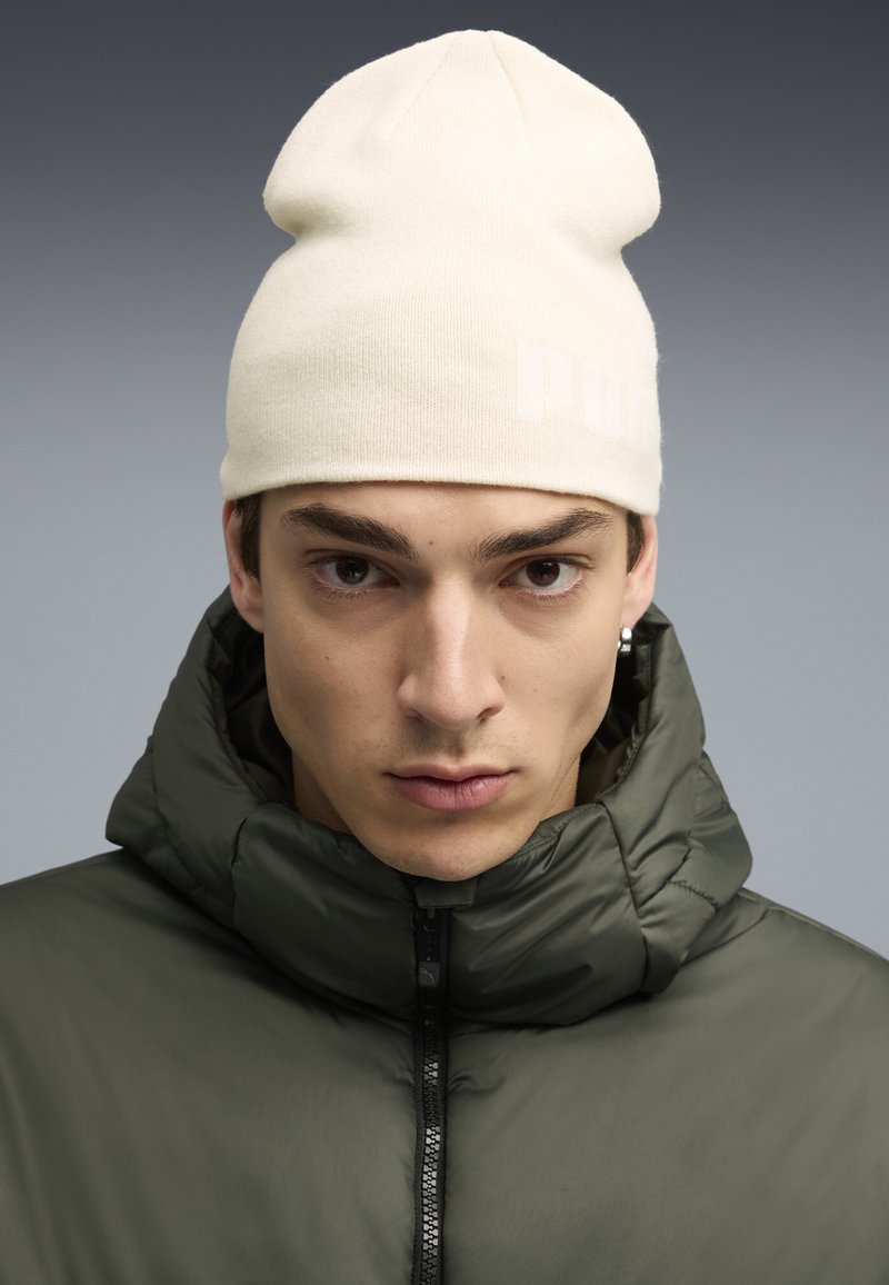 Cappello lavorato a maglia color crema con una vestibilità morbida, caratterizzato da un dettaglio del logo. Abbinato a una giacca a vento verde scuro, con zip frontale e colletto alto.