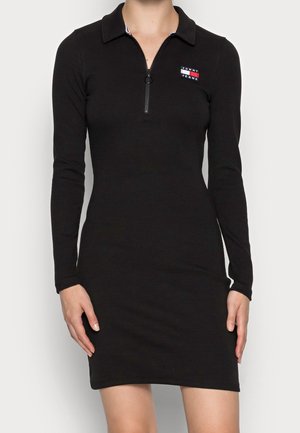 Femme portant une robe noire ajustée à manches longues avec un col, une fermeture éclair à l'avant et un petit logo Tommy Jeans sur la poitrine.