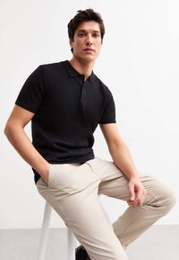 Zwarte polo met geribde structuur, drie knopen en een klassieke kraag, gecombineerd met beige broek. Man zit op een kruk.