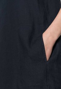 Ruka zasunutá do kapsy tmavě modré látky, ukazující texturu tkaniny a detaily šití na rukávu a těle.