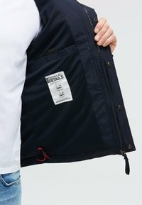 Granatowa kurtka z poliestrowym zewnętrzem i siateczkową podszewką. Posiada wewnętrzną kieszeń, zamek błyskawiczny oraz regulowany dół z czerwonymi akcentami.